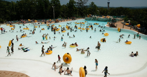 A busy water park with people swimming and floating on yellow inner tubes in a large pool surrounded by trees.