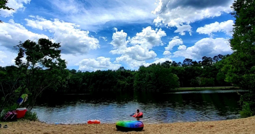 A person sits on a colorful inner tube by a sandy beach, with a calm lake and fluffy clouds in the background.