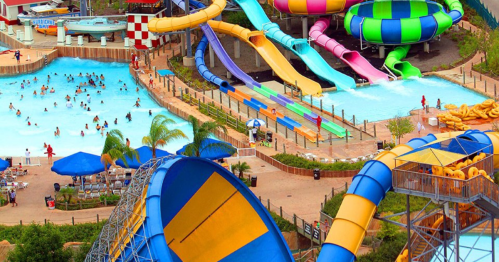 A vibrant water park featuring colorful slides, a wave pool, and lounging areas with umbrellas and palm trees.