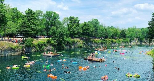 A lively swimming area with people on colorful floats, enjoying a sunny day by a green, tree-lined lake.