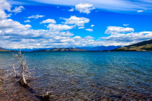 A serene lake with clear blue water, surrounded by mountains and a sky filled with fluffy clouds.