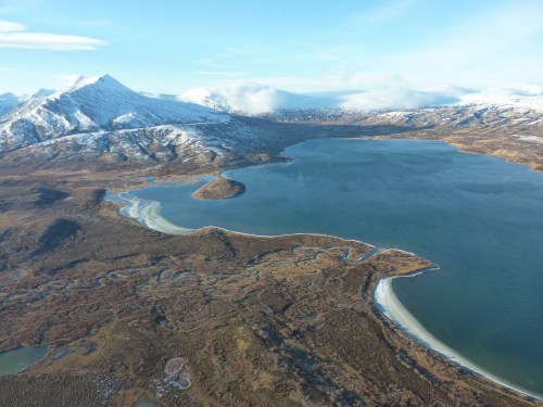 Aerial view of a serene lake surrounded by snow-capped mountains and lush, green landscapes under a clear blue sky.
