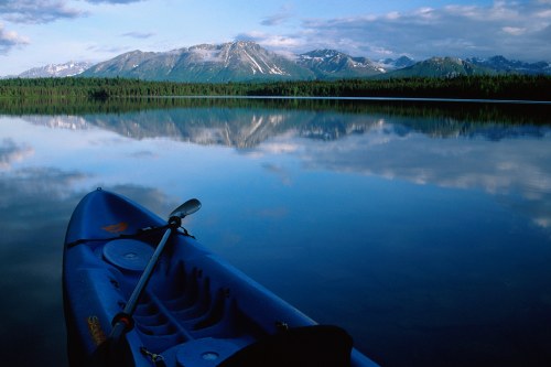A blue kayak rests on calm water, reflecting mountains and a cloudy sky in the background.