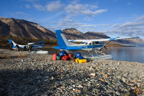 Two seaplanes parked by a rocky shore, with colorful backpacks and gear arranged nearby, under a clear blue sky.