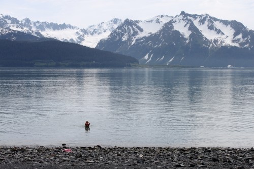 A person wades in calm water with snow-capped mountains in the background, creating a serene landscape.