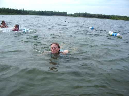 A girl swims in a lake with two children playing in the background, surrounded by trees and floating buoys.