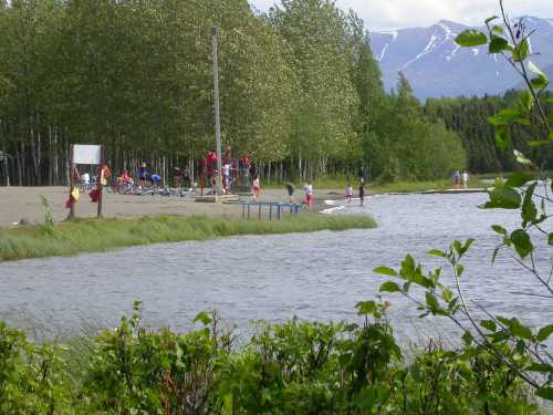 A sandy beach by a lake, with people enjoying activities and trees in the background, against a mountain backdrop.