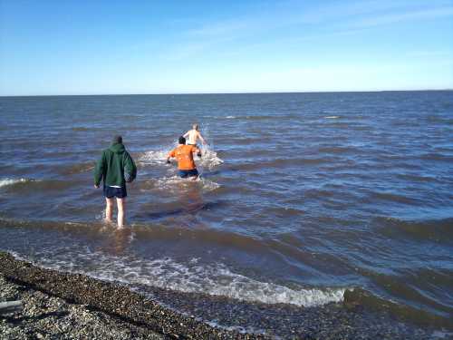 Three people wade into a calm lake, with one splashing water, under a clear blue sky.