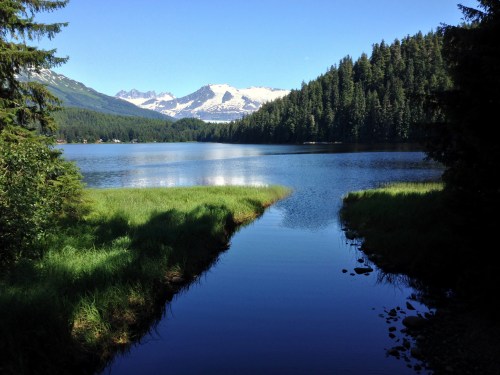 A serene lake surrounded by lush greenery and mountains, with snow-capped peaks in the background under a clear blue sky.
