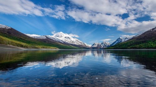 A serene mountain landscape with snow-capped peaks reflecting in a calm lake under a blue sky with scattered clouds.