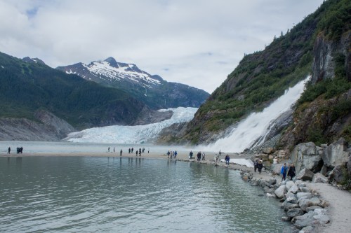 A scenic view of a glacier with a waterfall, surrounded by mountains and people walking along the shore.