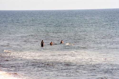 Three people wade in the ocean, surrounded by gentle waves under a clear sky.