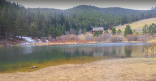 A serene lake surrounded by trees and mountains, with a cabin visible in the background and a grassy shore in the foreground.