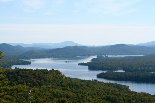 A serene landscape featuring a winding lake surrounded by lush green forests and distant mountains under a clear sky.