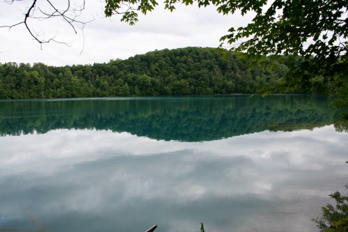 A serene lake reflecting lush green hills and cloudy skies, surrounded by trees.