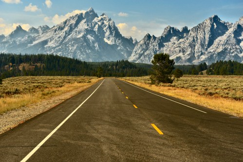 A straight road stretches through a grassy landscape, leading to snow-capped mountains under a blue sky.