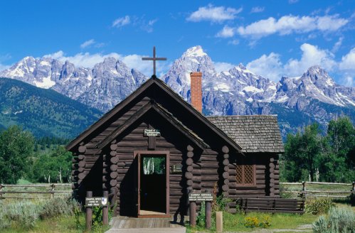 A rustic log chapel with a cross, set against a backdrop of majestic mountains and blue skies.