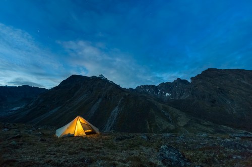 A tent glowing warmly at dusk, set against a backdrop of rugged mountains and a starry sky.