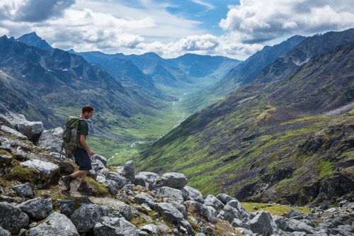 A hiker navigates rocky terrain in a lush valley surrounded by mountains under a partly cloudy sky.