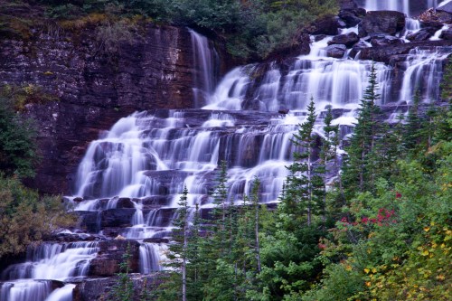 A cascading waterfall flows down rocky cliffs, surrounded by lush green trees and hints of autumn colors.