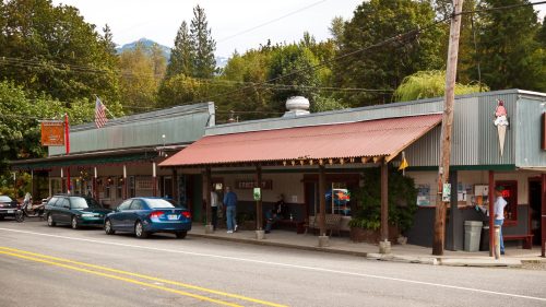 A small town street featuring shops with metal roofs, parked cars, and trees in the background under a clear sky.