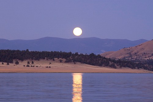A full moon rises over a calm lake, reflecting on the water with mountains in the background under a twilight sky.