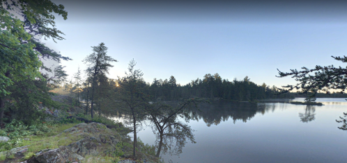 Serene lakeside view at dawn, surrounded by trees and rocky shoreline, reflecting calm water and soft morning light.