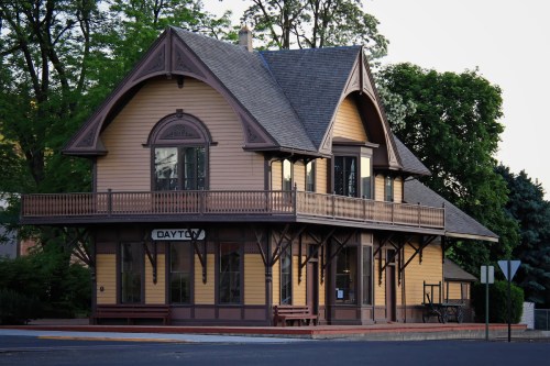 A charming, historic train station with a yellow exterior and a large balcony, surrounded by trees.