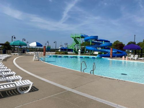 A sunny pool area featuring a water slide, lounge chairs, and umbrellas, with clear blue water and a blue sky.
