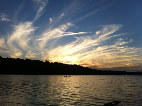 A serene sunset over a lake, with wispy clouds and two people kayaking in the foreground.