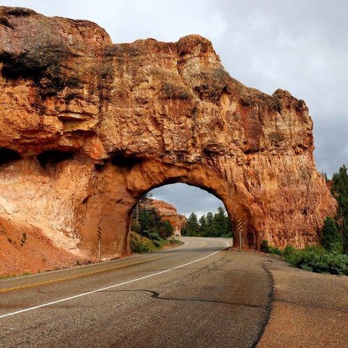 A scenic view of a natural rock arch over a winding road, surrounded by rugged cliffs and cloudy skies.