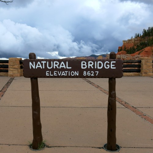 Sign for Natural Bridge at an elevation of 8,627 feet, with cloudy skies and rocky terrain in the background.