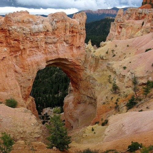 A natural rock arch stands amidst rugged terrain, surrounded by trees and distant mountains under a cloudy sky.