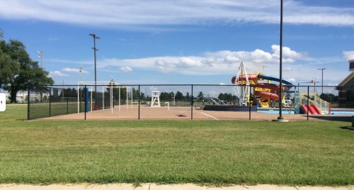 A fenced area with a soccer goal, a lifeguard stand, and a water slide near a pool under a blue sky.