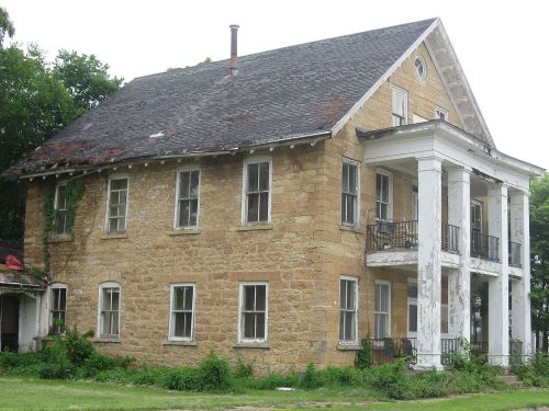 An old, two-story stone house with a sloped roof, white columns, and overgrown vegetation around it.