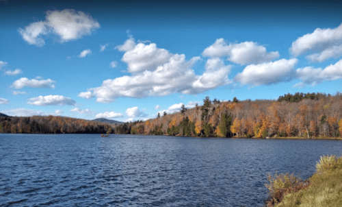 A serene lake surrounded by autumn trees under a blue sky with fluffy clouds.