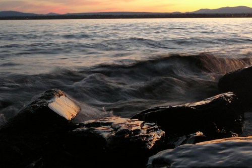 Waves gently lap against dark rocks at sunset, with a colorful sky and distant mountains in the background.