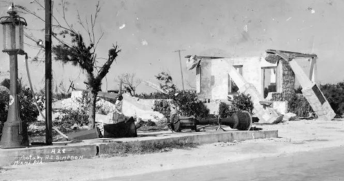 Black and white photo of a damaged building with debris, a lamp post, and trees in a desolate landscape.