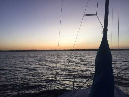 A serene sunset over calm waters, viewed from a sailboat with a blue sail and silhouetted rigging.