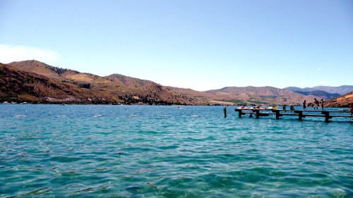 A serene lake with a wooden pier, surrounded by rolling hills and mountains under a clear blue sky.