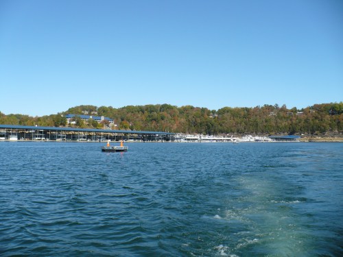A serene lake scene with a marina, boats, and colorful trees on the shore under a clear blue sky.