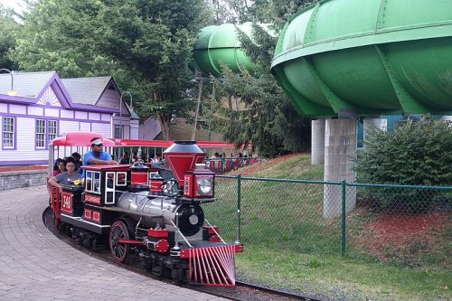 A small red train with passengers rides along a track near green water slides in a park setting.