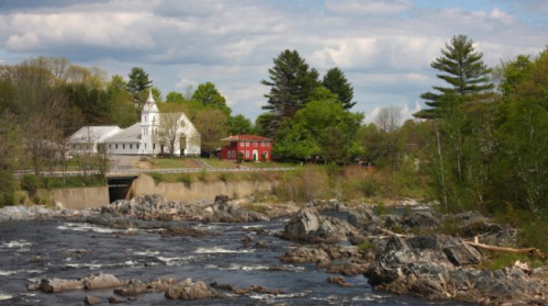 A scenic view of a river with rocky banks, featuring a white church and red building surrounded by greenery.