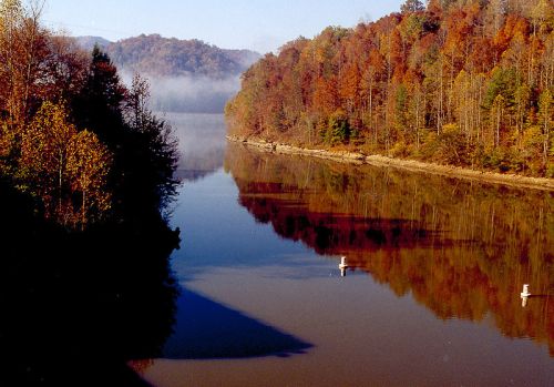 A serene river reflects colorful autumn trees and misty mountains under a clear sky.
