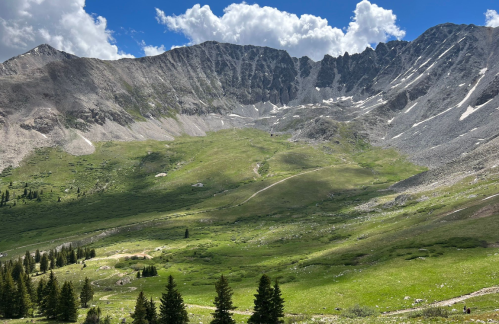 A scenic mountain landscape with green meadows, rocky peaks, and a blue sky dotted with clouds.