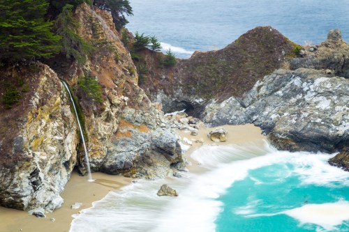 A scenic coastal view featuring a waterfall cascading onto a sandy beach, with waves crashing against rocky cliffs.