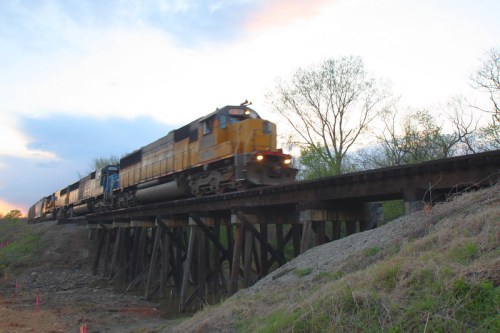 A yellow train speeds across a wooden trestle bridge at sunset, with trees and a cloudy sky in the background.