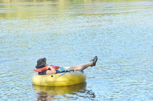 A person relaxes on a yellow inner tube in calm water, wearing a hat and life jacket.