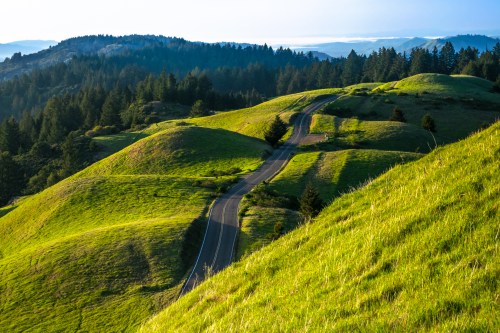 A winding road cuts through lush green hills under a clear blue sky, surrounded by distant mountains and trees.