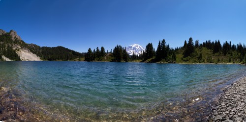 A serene lake surrounded by trees and mountains under a clear blue sky, with a snow-capped peak in the background.